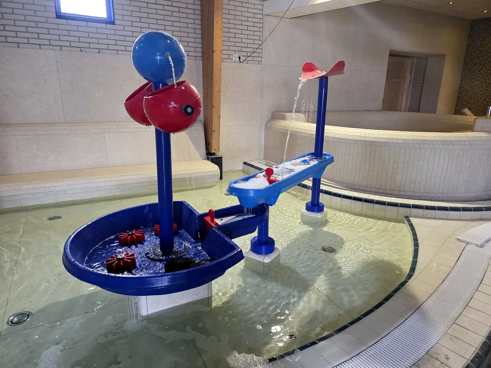 Blauw waterspeelelement met schalen, rode schoepen en een goot in het peuterbad van Paardekreek
